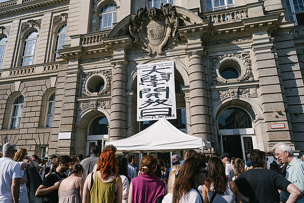 Im Bild ist der Eingang der Hardenbergstraße 33 mit dem diesjährigen Bannerdesign zu sehen. Menschen warten vorm Eingang. Es ist sonnig.